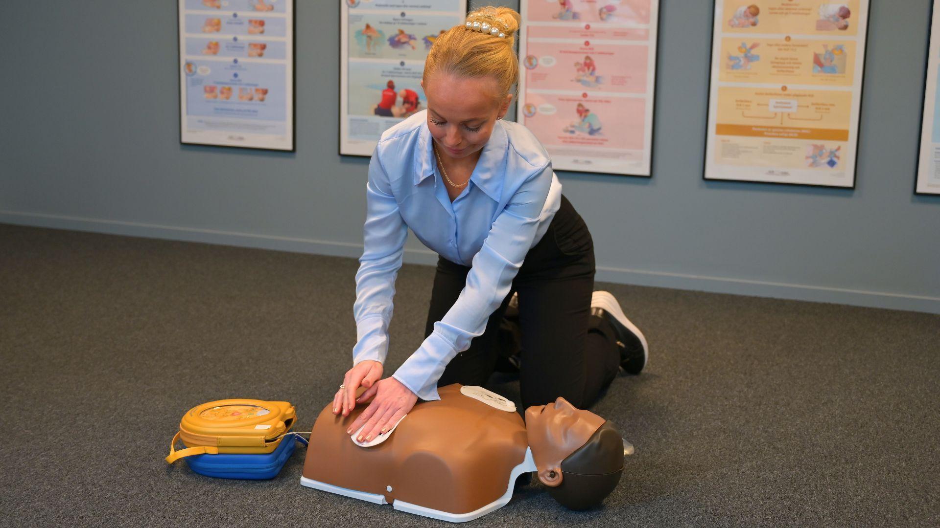 En kvinna sitter på knä på golvet och sätter elektroder från en hjärtstartare på en HLR-docka