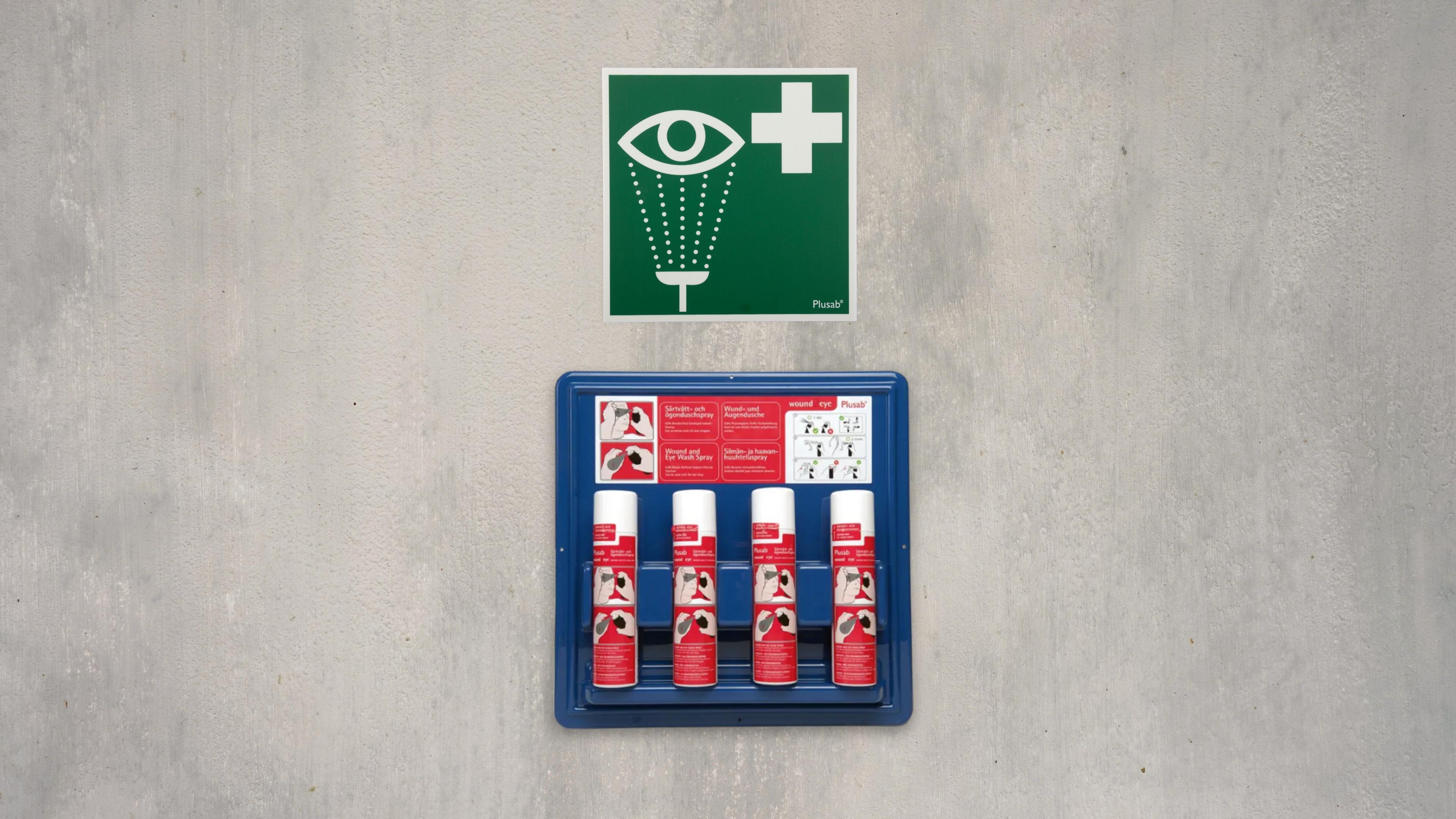 En blå hållare med fyra flaskor ögondusch sitter på en betongvägg. Ovanför sitter en grön skylt som indikerar ögondusch.