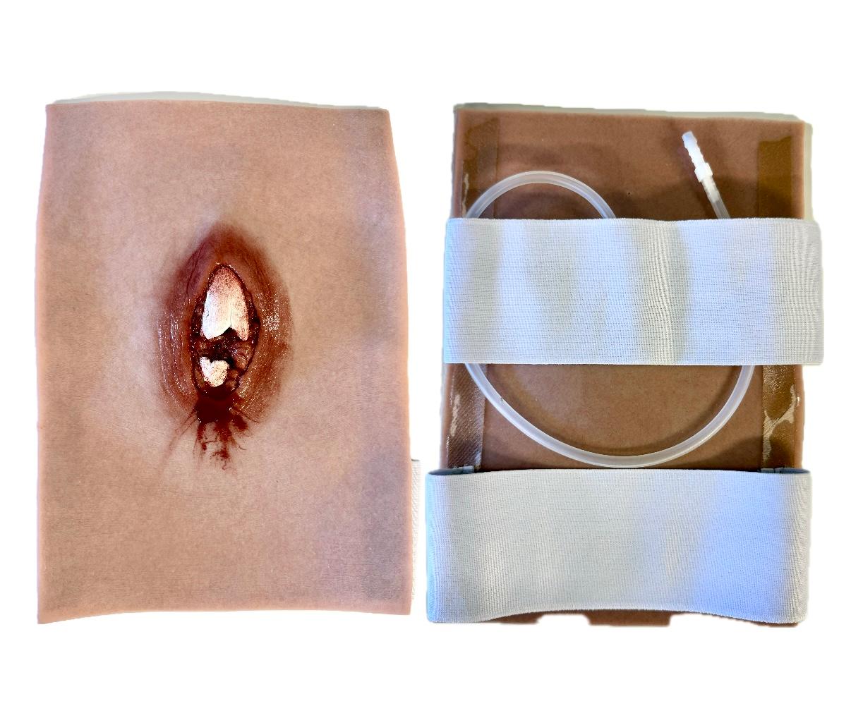 Rektangulär sårsimulator med ljus hud och ett sår med uppstickande benbit. På baksidan finns två remsor resår och en plastslang.